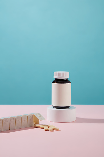 The tablets are emptied onto the table from a compartment of the daily pill box. An unlabeled medicine bottle is placed on a white podium. Blue pink background. Copy space.