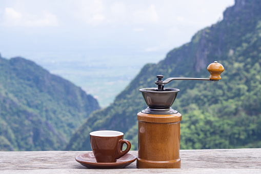 Manual coffee grinder and a coffee cup on wooden table with beautiful scenery view of mountains. Morning coffee feeling happy and freedom. Space for text.