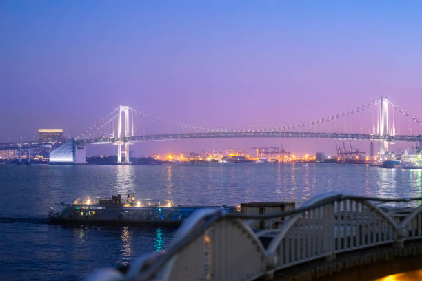 This image offers a picturesque view of Tokyo's Rainbow Bridge bathed in the soft glow of twilight. The photograph, taken in the evening in Tokyo, depicts the bridge's elegant suspension design against a backdrop of a pastel-colored sky that transitions from blue to a gentle pink. The ambient lighting of the bridge and surrounding buildings adds warmth to the scene, while the waters below reflect the calmness of the evening. This urban landscape captures the beauty of the city as it winds down, with the iconic bridge providing a connection not only across the bay but between the day and night.