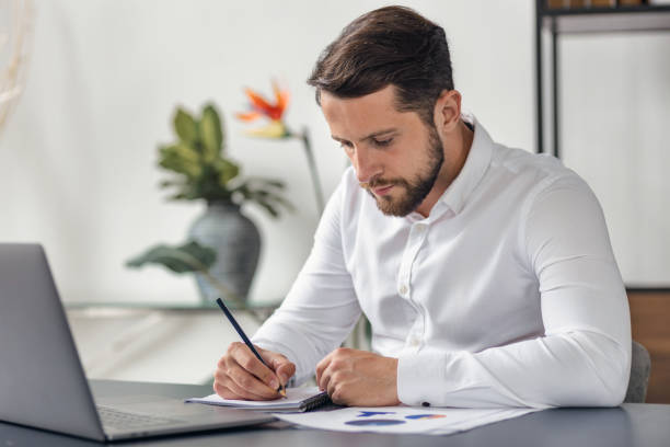 istock Concentrated man working using laptop takes notes in notepad sitting in office 1888823475