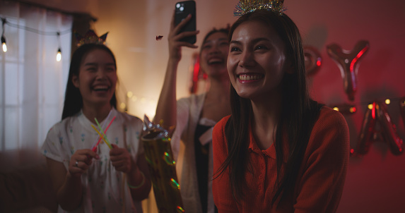 Selective focus, young asian woman and beautiful female friends wearing crown headband countdown and throwing confetti enjoying celebration together and use smartphone record video