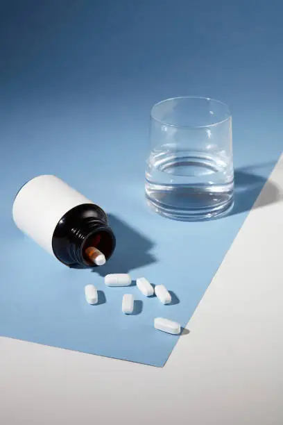 Close-up of white tablets poured onto a table from a white-label medicine bottle with a glass of water. New drug advertisement with delicate background.