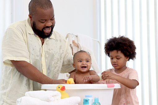 Happy African family, father bathing kid in tub, dad and son brother washing and cleaning little toddler girl daughter child in bath. Toddle baby infant has fun while taking bubble bath in bathroom.