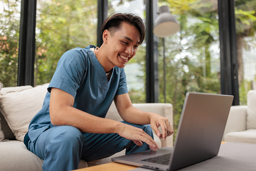 Young Asian man, dressed in a nurse uniform, is using laptop on the sofa in living room.