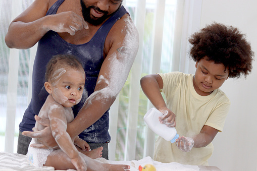 Happy African family, toddle baby infant sits on towel after takes a bath in bathroom, father and son brother hold dusting powder bottle and apply talcum powder on body of little kid daughter child.