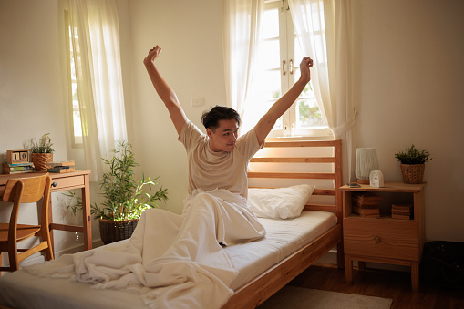 A young Asian man wakes up in bed in the morning and stretches.