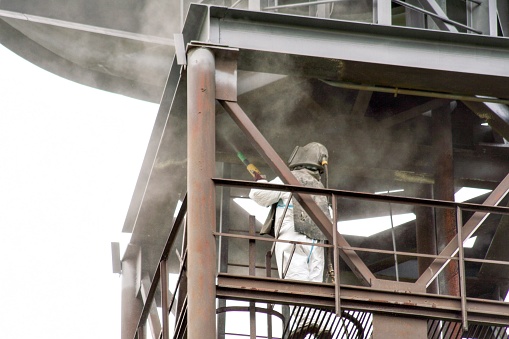 Sand blasting process. Industrial worker in protective uniform cleaning surface of big steel construction before painting on high altitude.