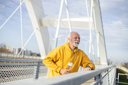 Active Senior man standing on the bridge teaking a break from a workout holding water bottle