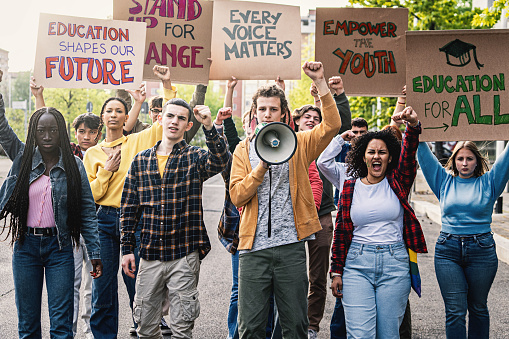 A diverse group of young students marching on the street, raising clenched fists and holding signs advocating for change in education.