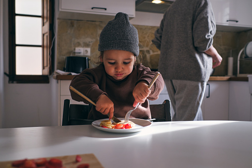 Cute little boy sitting at table and eating tasty food with fork and knife in kitchen with crop mother