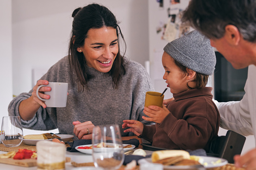 Charming child between smiling mother with cup of hot drink and crop unrecognizable father at kitchen table in morning