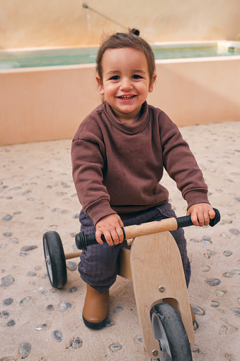 Charming child in casual apparel with toothy smile looking at camera while sitting on small bike outdoors