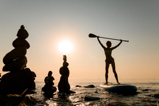 mujer delgada en traje de baño rema en la tabla de sup y sostiene el remo sobre su cabeza - paddleboard oar women lake fotografías e imágenes de stock