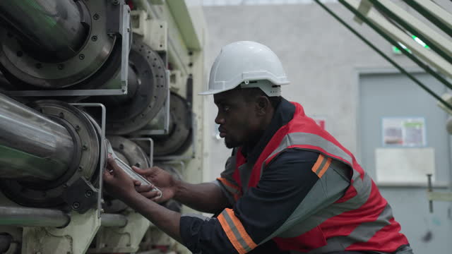 Close up shot of professional black male engineer with protective workwear adjusting the position of the roll of the manufacturing machine by using wrench. African male inspecting the machinery of the production line of paper factory.