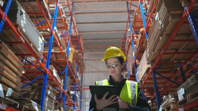 Mid adult white American female engineer inspector with protective workwear and safety vest standing and writing note on digital tablet after examining the cardboard products quality on a pallet in warehouse at a paper factory.