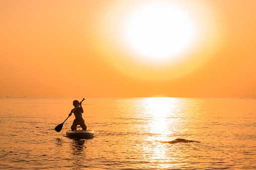 Girl wearing sun glasses and white bikini poses sitting on a sup board and oar using paddle. Sunset in the background.