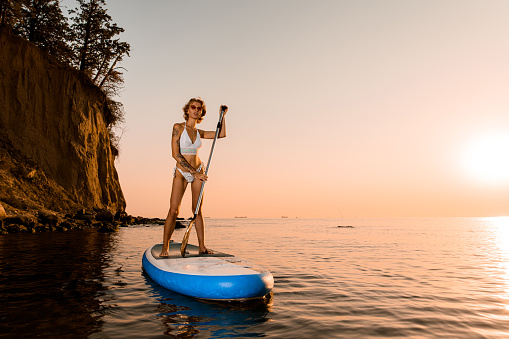 Beautiful woman traveler on stand up paddle board at quiet sea with bright sunset and rock cliff on background