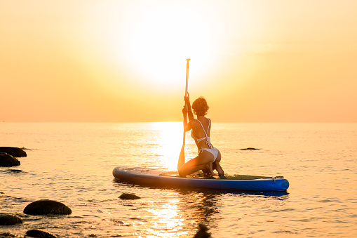 Active girl wearing white bikini poses sitting on sup board with paddle right against beautiful sunset