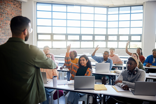 Diverse group of smiling college students raising their hands while asking their teacher questions during a lesson in a classroom at school