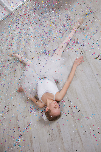 Girl in white ballet dress with ballet shoes lying on the floor in living room with confetti falling down on her.