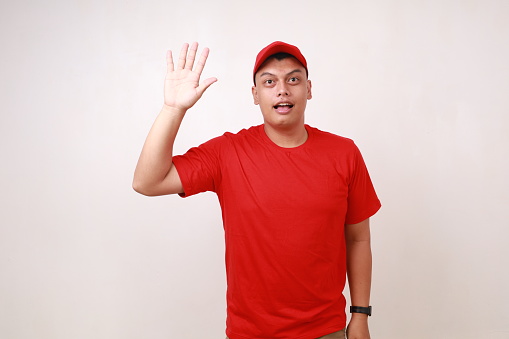 Handsome young asian courier raising waving his hand, saying hello, against white background