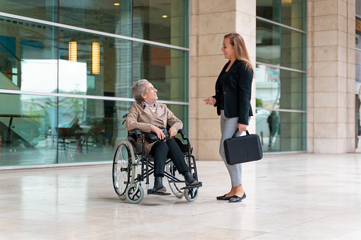Senior woman in wheelchair talking to friend or colleague outdoors in the city