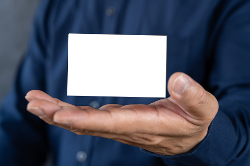 A man wears a blue jeans shirt and shows a blank business card. A horizontal mockup blurred background. A man holds a white business card on a concrete wall background.