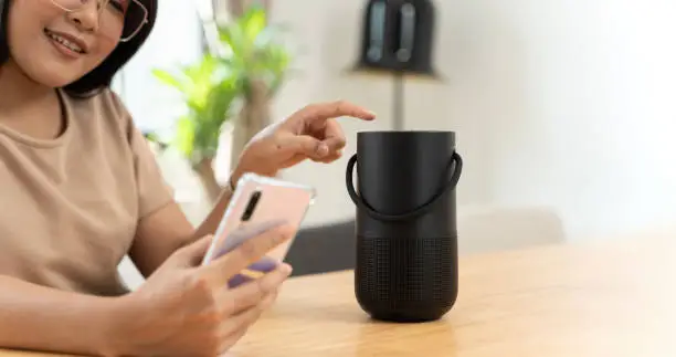 Photo of Woman streams music or podcasts to smart speaker from mobile phone at home.