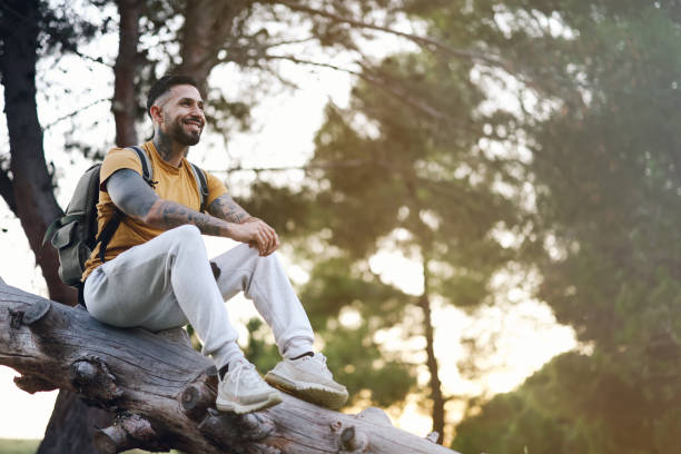 istock Smiling tattooed model man resting sitting on tree trunk in forest at sunset with back light 1889388315