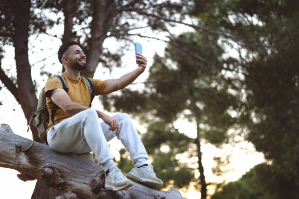hiker taking a selfie with his smartphone sitting on a tree trunk
