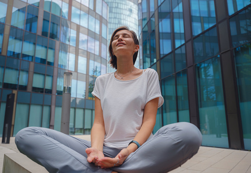 De-stress, unwind idea. peaceful female freelance employee practicing yoga exercises while sitting in bunch under glass skyscrapers after hard working day in office, holding hands in mudra