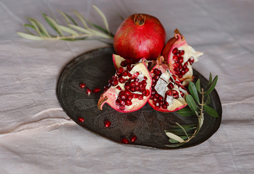 Image of Still Life with Turkish Pomegranate and olive branch on old retro plate. Dark wood background, antique copper plate. Fresh ripe whole pomegranates, opened pomegranate and seeds