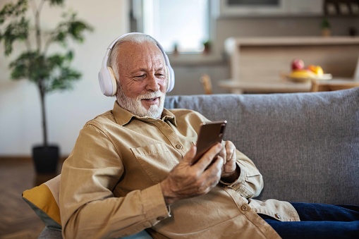 Senior man wearing headphones listens to favorite Mp3 digital music on smartphone at home while sitting on the bed in the living room, elderly man enjoys stereo high quality audio sound.