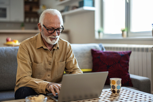 Smiling senior man using laptop at home. Handsome old man wearing eyeglasses working on laptop in living room. Portrait of elderly grandfather using computer.
