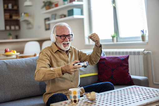 Bearded senior man with gamepad playing video game at home. Elderly man with gamepad playing video game in the living room during the day.