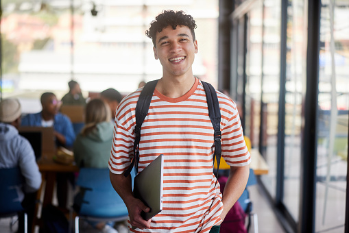 Portrait of a smiling young male college student standing with his laptop a backpack in a cafeteria at school