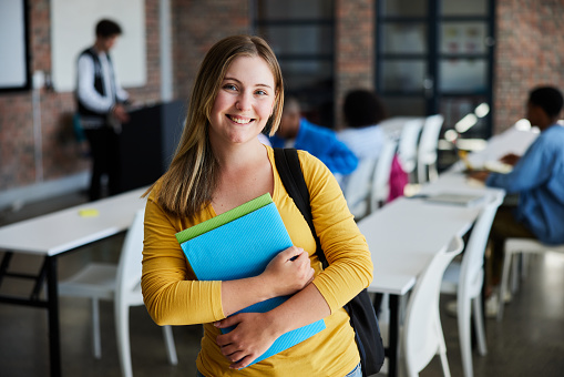 Portrait of a young female college student holding textbooks and smiling while standing in a classroom at school