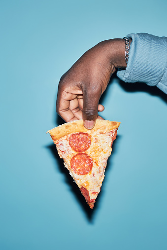 Vertical close up hot African American male hand holding slice of pepperoni pizza isolated on light blue background