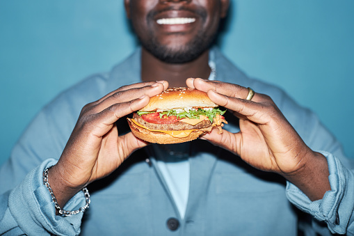 Close up shot of tasty meaty hamburger with vegetables in hands of Black man standing at light blue background
