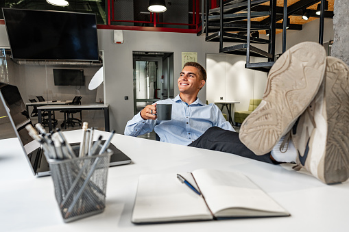 Smiling young businessman relaxing on chair with his feet resting on table in the office