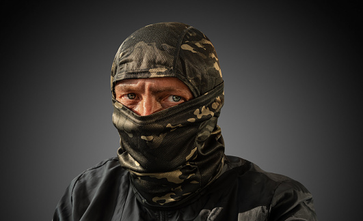 Military man with dirty face and camouflage mask. The concept of a person after military action or fraud, robbery. Close-up. A man on a dark background in a balaclava. Looking away. Fright.