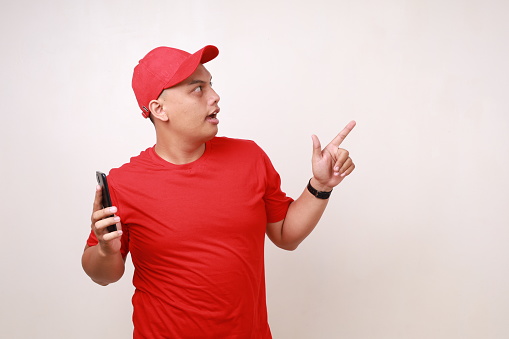 Shocked Asian delivery guy employee man in red standing while holding a cell phone and pointing sideways