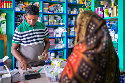 An Indian pharmacist in an Indian pharmacy attends to a customer sari woman who needs a medicine and caculate with the calculator the price of the drug