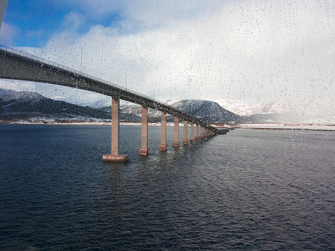 Low angle view of a bridge crossing the cold blue sea from a wet ferry window while raining, Lofoten Islands, Norway