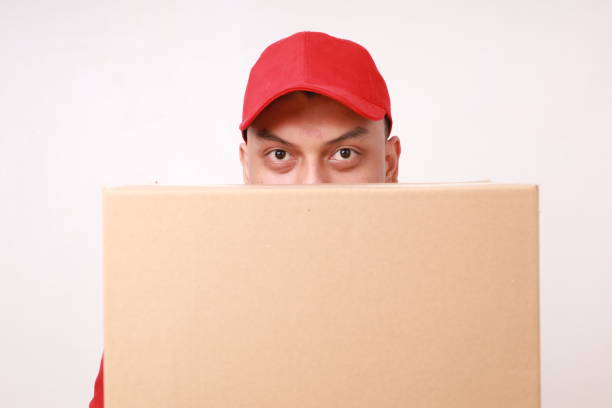 istock Asian courier man in red standing and hiding in cardboard box. Isolated on white 1889779112
