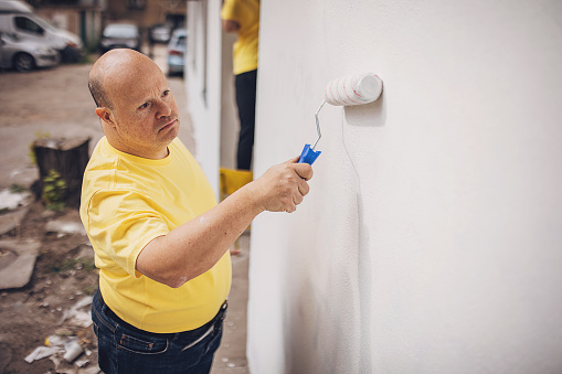 Man with down syndrome volunteer painting building wall outdoors.