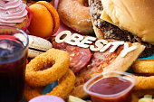 Full frame image of wooden letters spelling 'Obesity' on sweet and savoury junk foods, glass of iced cola, cheeseburger, glazed ring doughnut, macaron, cupcake with butter icing, pepperoni pizza slice, battered onion rings, French fries, tomato ketchup