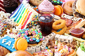 Full frame image of letter dice and wooden letters spelling out 'Fat','BMI',  'Unhealthy', 'Diet', 'Obesity' and 'Weight' on sweet junk foods, glass of iced cola, cheeseburger, battered onion rings, pizza slices, potato waffle, glazed ring doughnuts