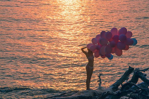 Young woman walking with many balloons in sunset at the ocean beach