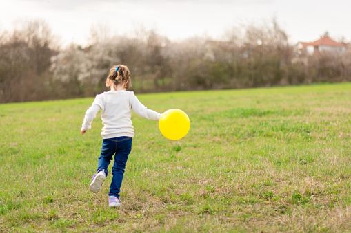 Rear view of a happy little girl is running across the meadow with a yellow balloon in her hand. Copy space